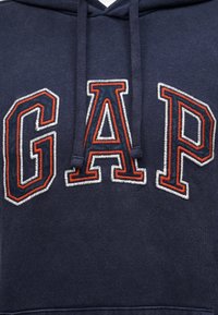 Μπλε φούτερ με κουκούλα σε ναυτικό μπλε, με έντονα κεντημένα γράμματα "GAP" σε ναυτικό μπλε, περιτυλιγμένα με πορτοκαλί και λευκή ραφή πάνω στο στήθος.