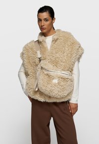 Chaleco beige de piel sintética con un acabado texturizado y esponjoso, combinado con un bolso bandolera a juego que cuenta con un bolsillo con cremallera y una correa ajustable.