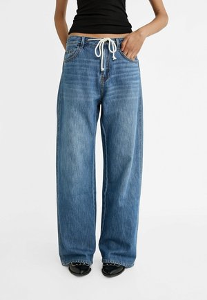 Osoba nosząca luźne niebieskie dżinsy z białym sznurkiem i czarne buty, ręka spoczywa w kieszeni, na jednolitym białym tle.