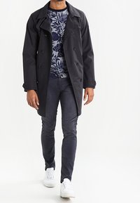 Mann läuft, trägt einen schwarzen Doppelreiher-Mantel über einem blauen Blumenmuster-Pullover, dunkle Hosen und weiße Turnschuhe vor einem weißen Hintergrund.