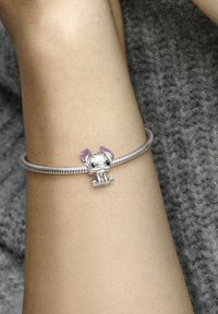 Bracciale d'argento con un ciondolo in stile cartone animato che presenta il volto di un personaggio e orecchie viola, posizionato su un braccio testurizzato contro uno sfondo grigio.
