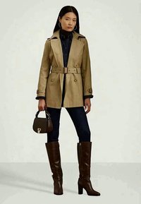 Γυναίκα με μπεζ παλτό με ζώνη, μαύρο παντελόνι, σκούρες καφέ μπότες έως το γόνατο, κρατώντας μια μικρή καφέ τσάντα, στέκεται σε απλό φόντο.