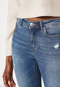 Jeans in denim azzurro chiaro con dettagli strappati, chiusura con bottone argentato e design a cinque tasche. Indossati con un maglione in maglia beige.