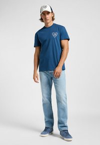 Mörkblå bomullst-shirt med hjärnsgrafik på bröstet, ihop med ljusblå jeans och en vit och mörkblå keps med rundad skärm.