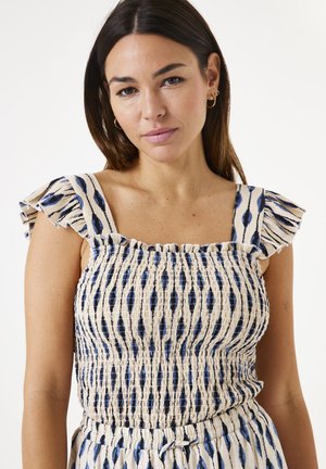 Haut à motif crème et marine avec des bretelles à volants, un corsage smocké et un tissu texturé. Présente des motifs de rayures horizontales et verticales.