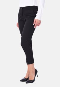 Pantaloni neri su misura con un design aderente, leggermente affusolati alle caviglie, tasche laterali e una texture liscia. Abbinati a tacchi neri a punta.