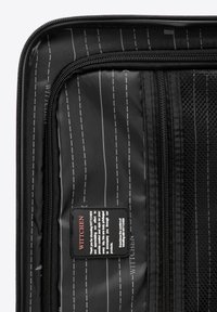 Intérieur d'une valise de voyage noire avec des poches en mesh, des compartiments zippés et une petite étiquette en tissu avec le nom de la marque. Motif à rayures horizontales.