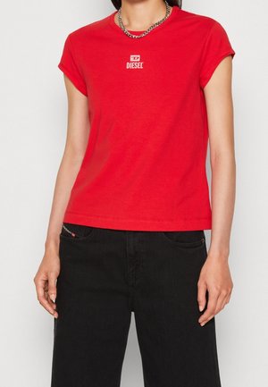 Kobieta w czerwonej koszulce Diesel z podwiniętymi rękawami i czarnych spodniach, ozdobiona srebrnym naszyjnikiem z łańcuszka.