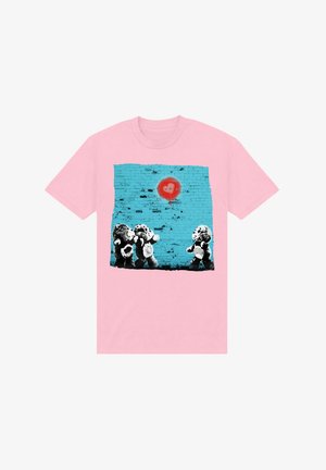 Růžové tričko s grafikou tří medvídků na pozadí tyrkysové cihlové zdi s červeným srdcem nad nimi.