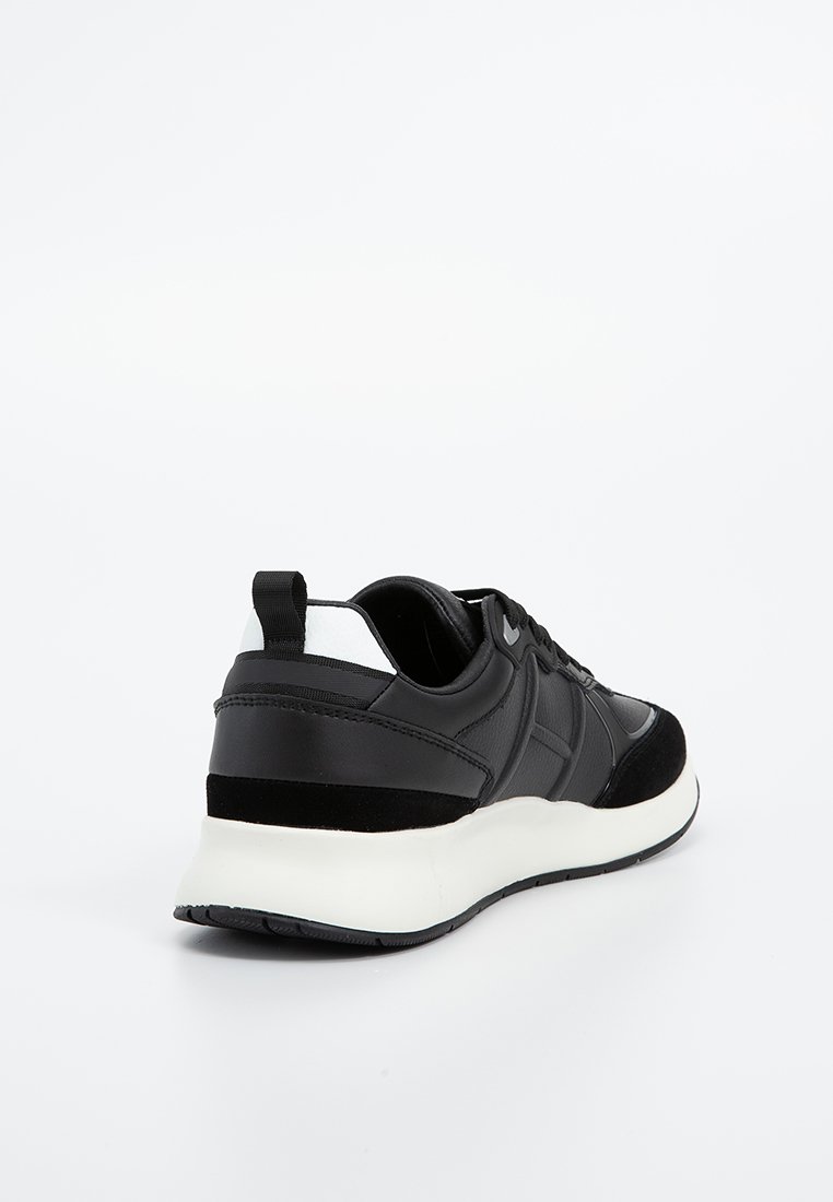 Baskets de sport noir et blanc avec une semelle blanche épaisse, une tirette noire et un dessus en cuir et tissu mélangés, montrées de l'arrière-gauche.