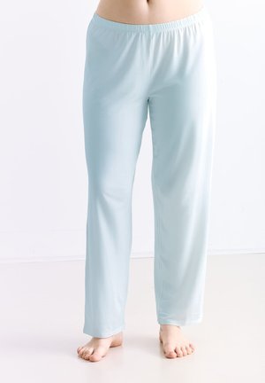 Person trägt hellblaue lockere Hosen mit elastischem Bund, steht barfuß auf einem glatten hellen Boden vor einem schlichten weißen Hintergrund.