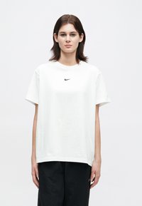 Fiatal személy, vállig érő barna hajjal, fehér Nike pólót és fekete nadrágot viselve, egy sima fehér háttér előtt áll.