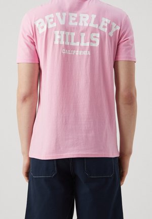 Person, die ein pinkes T-Shirt mit dem Text "Beverley Hills California" auf dem Rücken und dunkelblaue Hose trägt, von hinten gezeigt.