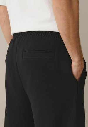 Nahaufnahme einer Person, die schwarze Hosen mit elastischem Bund und hinteren Leistentaschen sowie ein weißes Hemd trägt, Hand in der Seitentasche.