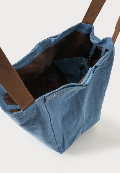 Sac cabas en tissu bleu avec des anses marron, ouverture en haut montrant la doublure intérieure sombre et une petite pochette intérieure zippée fixée avec un clip.