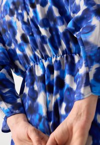 Mains jointes dans le dos portant une robe à manches longues à motifs bleu, blanc et noir avec un détail froncé à la taille.