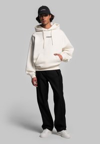 Felpa bianca con tasca frontale e cordini, logo nero sul petto, abbinata a pantaloni neri a gamba larga e sneakers bianche, indossata da un modello.