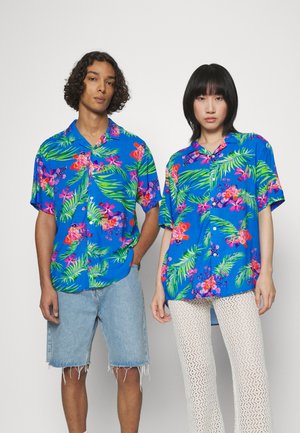 Blue short-sleeve shirts feature a tropical floral pattern with pink and green accents. One model wears denim shorts, the other white textured pants.