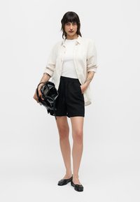 Γυναίκα όρθια με λευκό πουκάμισο πάνω από λευκή μπλούζα, μαύρες σορτς, μαύρα φλατ παπούτσια, κρατώντας μια μαύρη τσάντα, με τατουάζ στα χέρια.