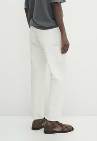Pantaloni cargo bianchi con una vestibilità rilassata, dotati di una tasca laterale e indossati con sandali in pelle marrone. Lo sfondo è semplice e minimale.