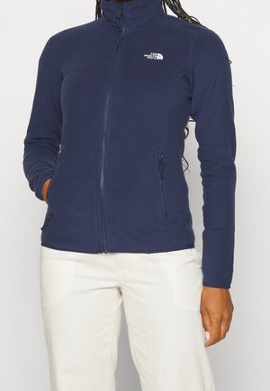 Kobieta w granatowej polarowej kurtce zapinanej na zamek błyskawiczny z logo The North Face na piersi i białych spodniach, ręce w kieszeniach.