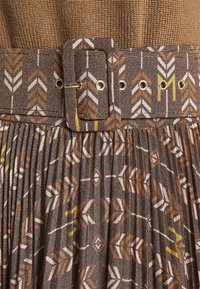 Jupe plissée marron avec des motifs géométriques en forme de flèches beige, blanc et jaune, accompagnée d'une ceinture en tissu assortie avec des œillets métalliques et une boucle rectangulaire.