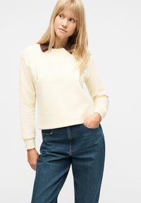 Ξανθιά γυναίκα που φοράει κρεμ φούτερ με ανάγλυφο το "PARIS" και μπλε τζιν, στέκεται με το ένα χέρι στην τσέπη, μπροστά σε λευκό φόντο.