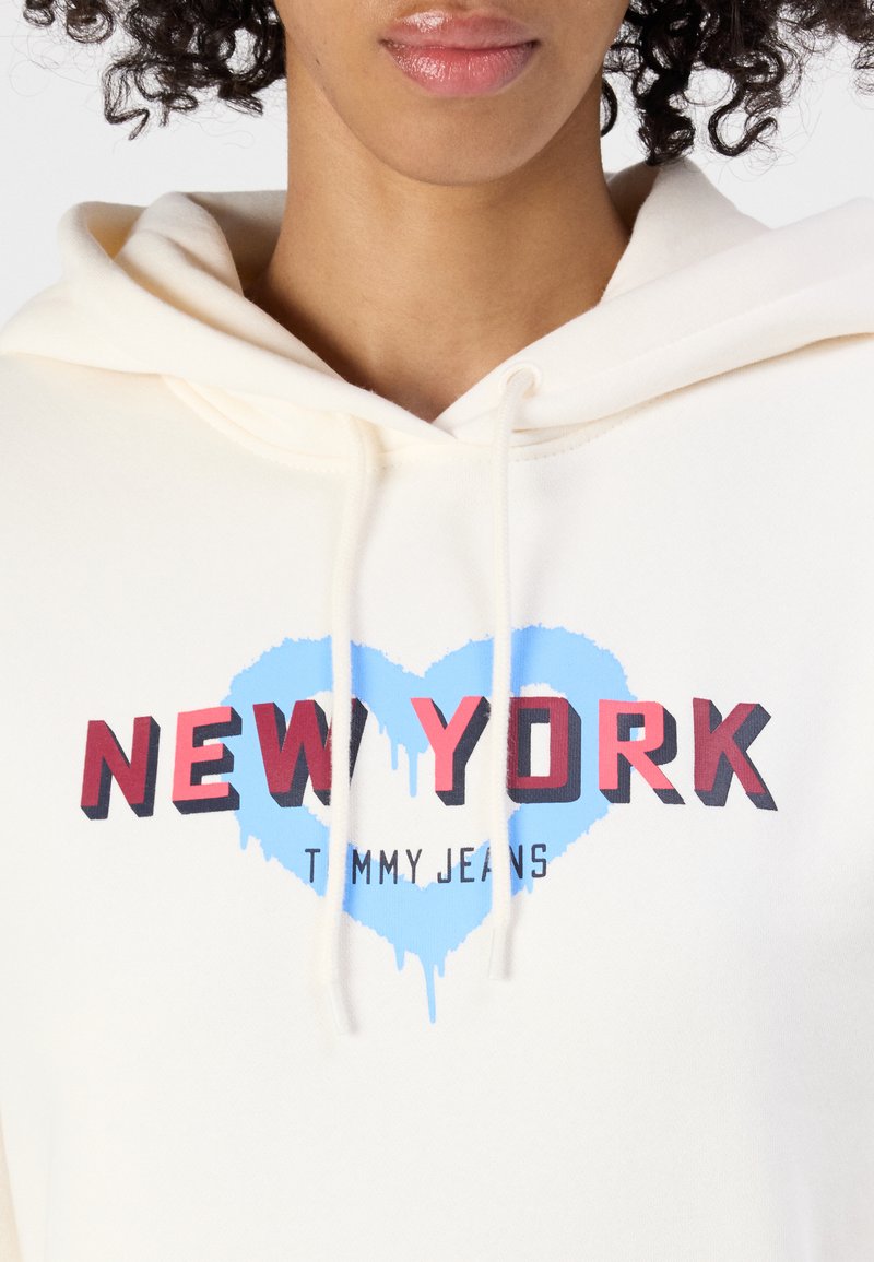 Helle cremefarbene Kapuzenjacke mit Kordelzügen, versehen mit einem blauen Herz-Graphic und rotem "NEW YORK"-Text. Der Stoff hat eine weiche Textur und ein lässiges Design.