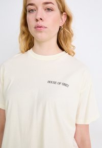 Crèmekleurig T-shirt van zachte stof, met een kleine zwarte tekstprint "HOUSE OF OBEY" op de linkerborst. Ronde halslijn.