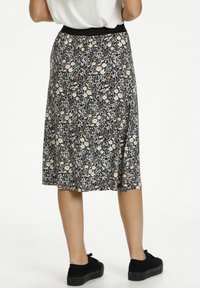 Jupe midi fleurie avec un fond noir, ornée de fleurs blanches et jaunes, une ceinture élastique, portée avec des chaussures noires.