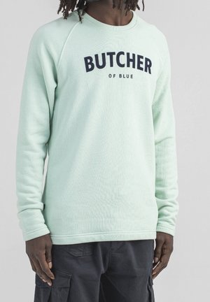 Mężczyzna w jasnozielonej bluzie z napisem "BUTCHER OF BLUE", ubrany w ciemne spodnie typu cargo, stoi na tle jednobarwnym.
