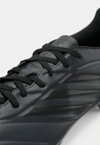 Μαύρο δερμάτινο ποδοσφαιρικό παπούτσι με μαύρα κορδόνια και διακριτικές ρίγες της μάρκας στο πλάι, εμφανιζόμενο σε κοντινή λεπτομέρεια.