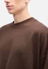 Sweatshirt marron à col rond, fabriqué en tissu doux. Doté d'une broderie subtile "MN" sur la poitrine. Texture lisse et coupe décontractée.