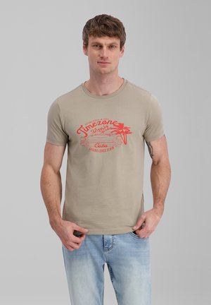 Mann trägt ein beiges T-Shirt mit rotem Vintage-Auto- und Palmen-Design, hellblaue Jeans, steht vor einem schlichten grauen Hintergrund.