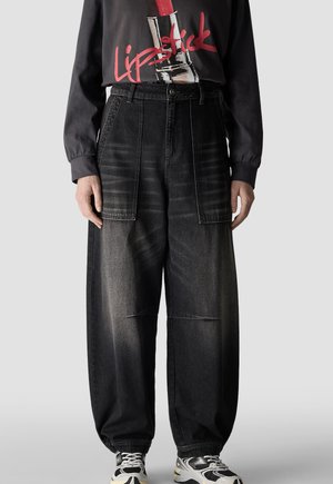 Person trägt lockere schwarze Jeanshosen mit großen aufgesetzten Fronttaschen, ein schwarzes Hemd mit rotem "Lipstick" Text und Grafik sowie schwarz-weiße Turnschuhe.
