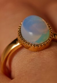 Gouden ring met een ovale cabochon opaal in blauw en groene tinten, gezet in een gestructureerde zetting. Gladde band met gegraveerde details.