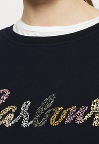 Tmavě modrý svetr s kulatým výstřihem. Obsahuje vícoloré květinové vyšívání v pastelových odstínech, které ukazuje různé vzory listů.