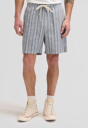 Mężczyzna noszący szare i białe szorty ze sznurkiem w pionowe paski, białe skarpetki oraz beżowe sneakersy za kostkę na jednolitym tle.