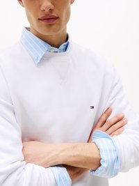 Sweatshirt branco com um pequeno logótipo, sobreposto a uma camisa listrada em azul claro e branco com punhos de colarinho abotoados.
