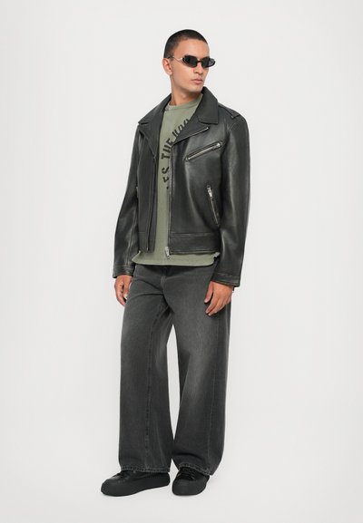 Μαύρο δερμάτινο μπουφάν μοτοσικλέτας, πράσινο γραφικό t-shirt, φαρδιά σκούρα τζιν, μαύρα παπούτσια και μικρές μαύρες γυαλιά ηλίου. Λείο υλικό και χαλαρή εφαρμογή.
