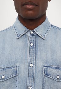 Camisa de mezclilla azul claro con cuello puntiagudo, cierre frontal con botones y dos bolsillos en el pecho con solapas. Textura suave, diseño informal.