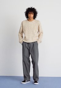 Suéter de punto de cable beige, pantalones grises de rayas sueltos y zapatillas negras. El fondo liso enfatiza los detalles de la ropa.