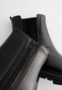 Bottines en cuir verni noir avec une finition texturée, dotées d'une fermeture éclair sur le côté et d'un panneau élastique. Détails cousus et semelle en caoutchouc.