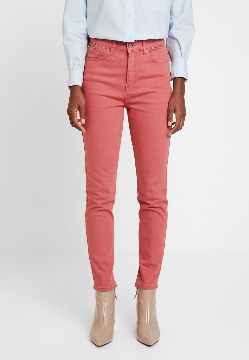 Jeans skinny rosas, mezcla de algodón, diseño de cintura alta, estilo de cinco bolsillos, combinados con botas de tobillo brillantes beiges, vista frontal sobre un fondo blanco.