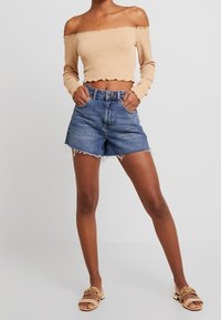 Béžový krátký top bez ramínek s texturovanou tkaninou, v kombinaci s modrými denimi šortkami s roztrhanými lemy. Nosené s hnědými plochými sandály.
