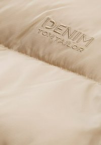 Bēša pufferkurta materiāls ar gludu tekstūru. Iekļauj izšuvumu "DENIM TOM TAILOR" logotipu kontrastējošā krāsā.