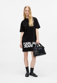 Crna oversized majica haljina s natpisom "KARL LAGERFELD JEANS" u bijeloj boji na rubu, u kombinaciji s crnom torbicom i crnim čarapama sa sandalama.