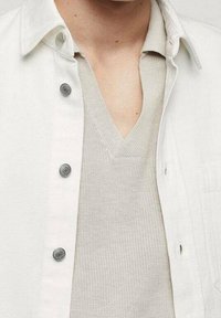 Camicia bianca con bottoni a vista sopra un maglione beige chiaro a V con tessitura a rilievo. Presenta bottoni argento e una vestibilità rilassata.