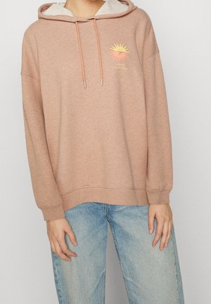 Personne portant un sweat à capuche marron clair avec un motif soleil et un jean bleu, debout devant un fond clair uni.