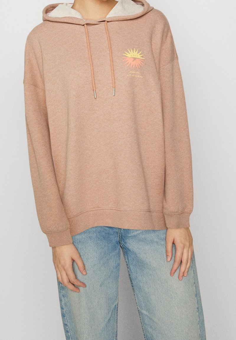 Personne portant un sweat à capuche marron clair avec un motif soleil et un jean bleu, debout devant un fond clair uni.